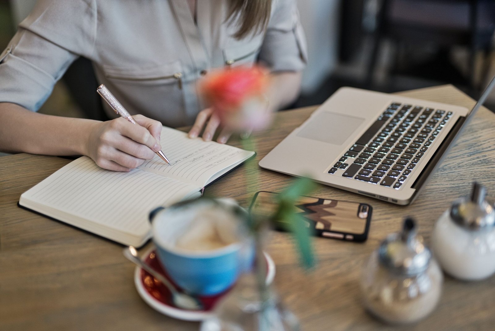 A person writing preparation notes in a notebook beside a laptop and coffee cup on a desk