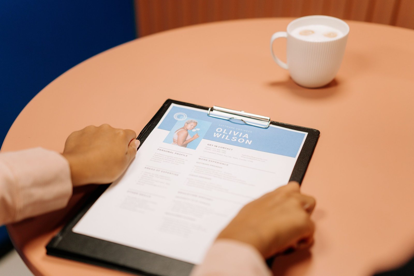 A hiring manager's hands holding a printed resume across a desk during an interview, coffee cup beside a laptop — the human review stage that follows the automated one
